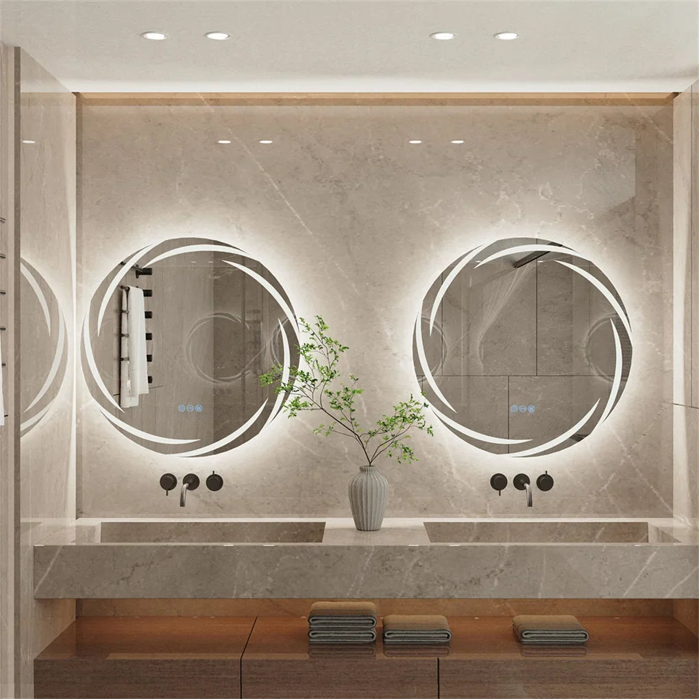 A modern bathroom interior with two large oval backlit mirrors mounted on a light stone wall above a double sink. The scene includes a vase with greenery, folded towels, and recessed ceiling lighting.